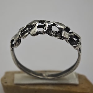 May include: A silver ring featuring an irregular, organic, molten-like band design with open cutouts and a textured, oxidised finish. The metal surface displays a series of raised, rounded shapes and hollow spaces that create a continuous, uneven pattern.