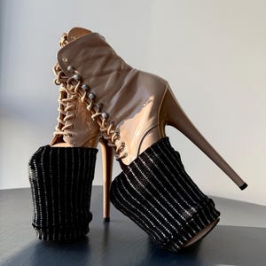 Puede incluir: Un par de botines de charol beige con tacones altos y detalles de cordones. Los botines tienen tacón de aguja y se combinan con calentadores de piernas a rayas negras y plateadas. Los botines están sobre una superficie azul oscuro.