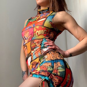 May include: A sleeveless, high-neck mini dress featuring a vibrant comic book print in red, yellow, blue, and orange. The dress has a ruched side detail with drawstring ties. The overall design is a bold and eye-catching statement piece.