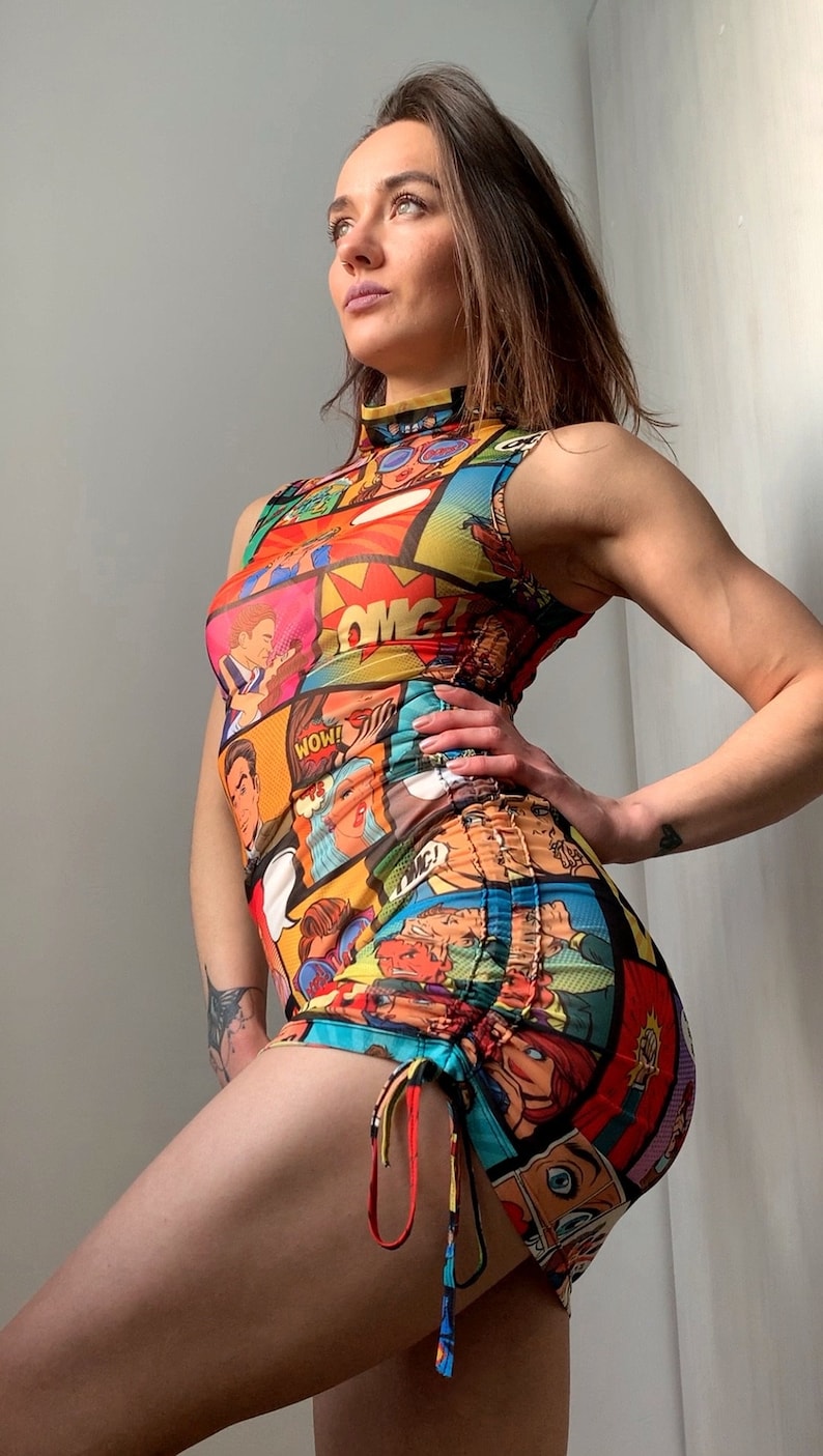 May include: A sleeveless, high-neck mini dress featuring a vibrant comic book print in red, yellow, blue, and orange. The dress has a ruched side detail with drawstring ties. The overall design is a bold and eye-catching statement piece.
