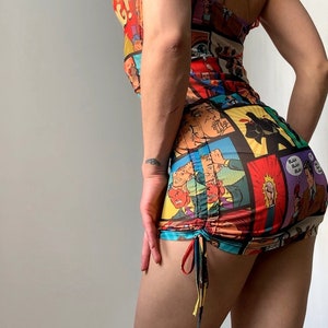May include: A colorful, sleeveless mini dress featuring a comic book print. The dress has a scoop neckline and a ruched skirt with drawstring ties. The print includes panels with speech bubbles and action scenes.