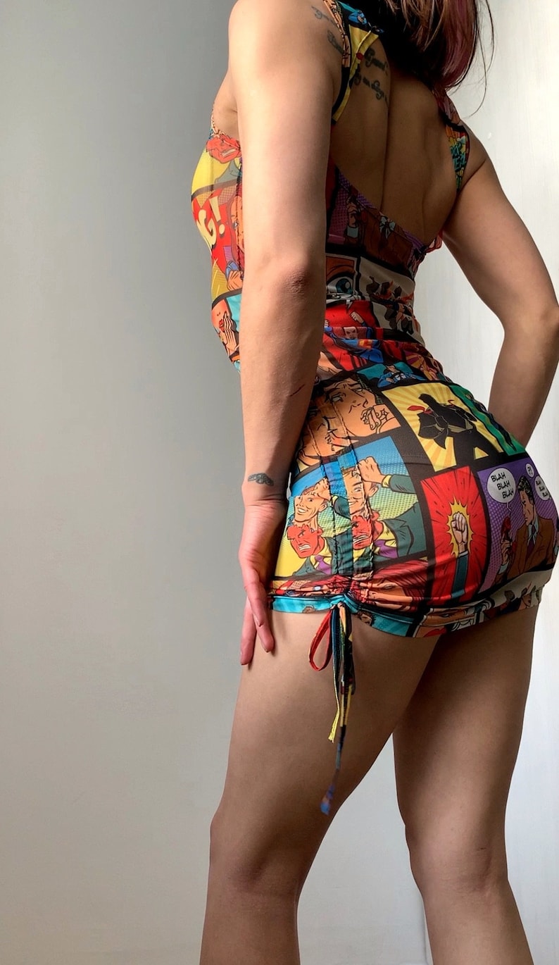 May include: A colorful, sleeveless mini dress featuring a comic book print. The dress has a scoop neckline and a ruched skirt with drawstring ties. The print includes panels with speech bubbles and action scenes.