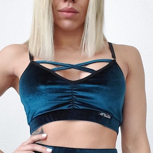 Op de afbeelding: Een tealblauwe sportbeha van fluweel met zwarte bandjes en een gekruist ontwerp. De beha heeft een zwarte elastische band aan de onderkant met de tekst "ATM STUDIO" erop gedrukt.