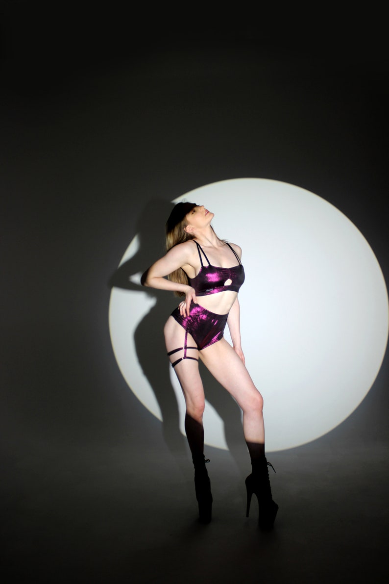 May include: A two-piece set of lingerie in a shimmering purple fabric. The set includes a crop top with thin straps and high-waisted shorts with garter straps. The model is wearing black platform heels. The background is a white spotlight.