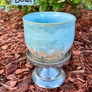 May include: A light blue ceramic bowl with a smooth, rounded shape and a subtle gradient transition to a tan base. The bowl is positioned on a silver-colored metal pedestal stand with a scalloped edge. The text "Bowl" is visible in the upper left.