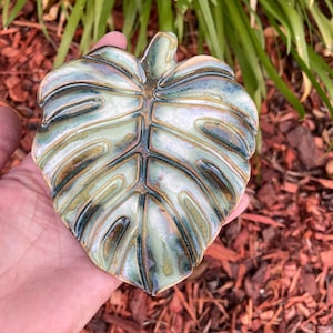 May include: A ceramic dish shaped like a monstera leaf with a glossy finish. The surface features a blend of sage green, deep forest green, and iridescent brown tones, with raised ridges defining the leaf veins and a smooth, contoured texture.