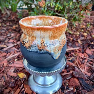 May include: A ceramic mug with a matte black base and a glossy, dripped glaze finish in shades of orange, cream, and dark blue. The mug features a wide, rounded body and a flared rim, resting on a silver-toned, scalloped pedestal stand.