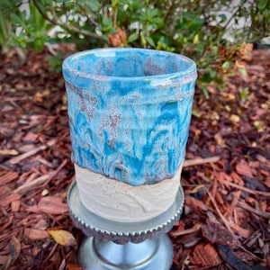 May include: A cylindrical ceramic tumbler featuring a two-tone design with a textured, off-white base and a glossy, mottled blue glaze on the upper portion. The vessel sits atop a small, silver-colored metal pedestal stand.