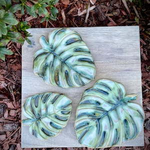 May include: Three ceramic dishes shaped like monstera leaves, featuring a glossy glaze in shades of light green, cream, and dark blue. The leaves have detailed, raised veins and organic, irregular edges, arranged on a square wooden board.