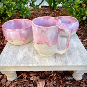 May include: A ceramic mug and two matching bowls featuring a speckled cream-colored base and a glossy pink glaze that drips down from the rim. The pottery has a rounded shape with a thick handle on the mug, showcasing a smooth, hand-crafted finish.
