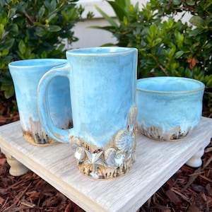May include: A set of three ceramic pieces including a tall mug with a handle, a smaller cup, and a bowl. Each piece features a light blue glaze on the upper portion and a textured, sandy-colored base decorated with sculpted seashells and starfish.