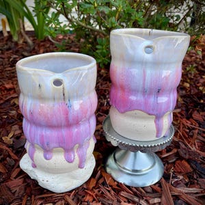 May include: Two ceramic pitchers with a cylindrical, ribbed body and a small circular hole near the rim. The glaze features a white base with dripping pink and purple hues. One pitcher rests on a silver-colored pedestal stand.