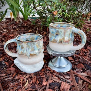 May include: Two ceramic mugs featuring a speckled cream base and a glossy, multi-colored drip glaze in shades of blue, tan, and brown. One mug rests on a white stone, while the other is elevated on a small, silver-colored metal pedestal stand.