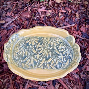 May include: An oval ceramic tray with a scalloped edge, featuring a glossy mustard yellow border and a recessed center decorated with a relief pattern of dark blue-grey coneflower blossoms and leaves across the entire surface.