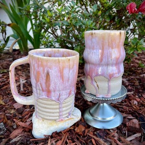 May include: A ceramic mug with a handle and a matching cylindrical cup, both featuring a textured beige base and a glossy, dripping pink and white glaze. The mug rests on a stone, while the cup sits on a small, silver-colored metal pedestal.