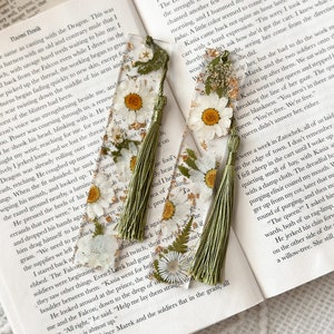 A picture showing two bookmarks. The bookmarks are made from clear resin and have white daisy flowers, green leaves, and gold flakes. The bookmarks come with a sage green tassel. They are aesthetic.