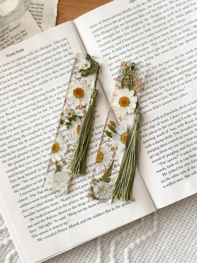 A picture showing two bookmarks. The bookmarks are made from clear resin and have white daisy flowers, green leaves, and gold flakes. The bookmarks come with a sage green tassel. They are aesthetic.