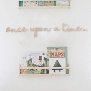 May include: A white wooden shelf with books and a wooden sign that says "once upon a time..." above it. There is a white armchair with a white pillow in front of the shelf. A wooden table with a basket of stuffed animals, a rainbow wooden arch, and a toy truck is in front of the armchair.