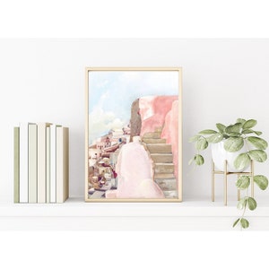 May include: A framed watercolor print depicting a Mediterranean coastal scene with pink buildings, stone stairs, and a blue sky. The artwork is displayed in a thin gold frame, positioned between a row of books and a potted plant on a white shelf.