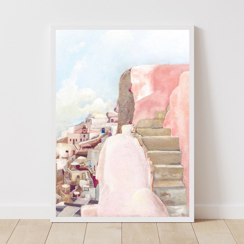 May include: A vertical watercolor painting in a white frame depicting a Mediterranean coastal scene with light pink walls, stone steps, and a cluster of white buildings under a pale blue sky. The artwork features soft pastel tones and architectural details.