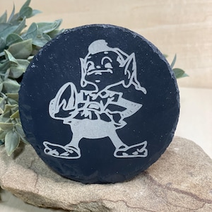 May include: A round, black slate coaster featuring a white etched image of a leprechaun-like character holding a football. The coaster is set on a light brown stone, with green foliage in the background. A decorative item.