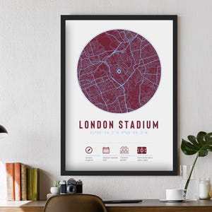 Puede incluir: Impresión enmarcada del estadio de Londres, con un mapa circular en burdeos y azul. El texto "LONDON STADIUM" está debajo, con coordenadas e iconos. La impresión se muestra en una pared blanca.