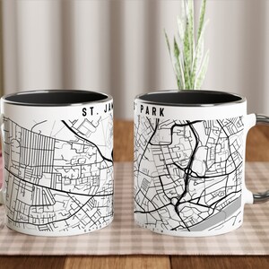 May include: Two white ceramic mugs with black rims. The mugs feature black and white maps of St. James Park and a city. The mugs are on a wooden table.