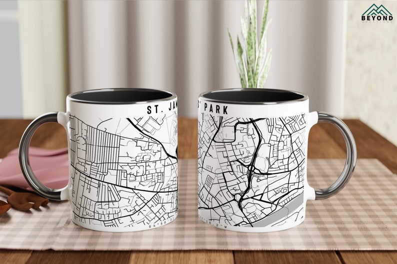 May include: Two white ceramic mugs with black rims. The mugs feature black and white maps of St. James Park and a city. The mugs are on a wooden table.
