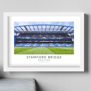 May include: A white framed print of Stamford Bridge, the home of Chelsea Football Club. The image shows the stadium's exterior with a blue and white color scheme. The text "Stamford Bridge, Chelsea, Opened in 1905" is printed below the image.