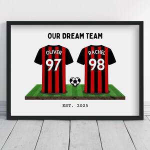 Puede incluir: Impresión enmarcada con dos camisetas de fútbol negras con rayas rojas, con los nombres "Oliver" y "Rachel" y los números 97 y 98. Un balón de fútbol y el texto "OUR DREAM TEAM" y "EST. 2025" también son visibles.