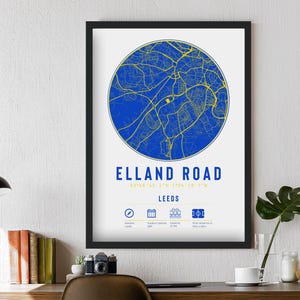 Puede incluir: Impresión enmarcada con un mapa azul y amarillo de Elland Road, Leeds. Incluye el texto "ELLAND ROAD" y coordenadas. Iconos detallan información del estadio. Se muestra en una pared blanca sobre un escritorio de madera.