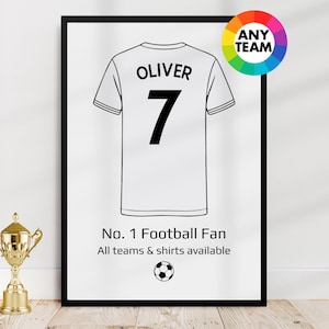 May include: Framed print featuring a white football jersey design with the name "OLIVER" and the number "7" in black. The print includes the text "No. 1 Football Fan" and a soccer ball graphic. A gold trophy is in the foreground.