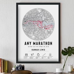 May include: A framed print with a black border. The print features a circular map with a red route, labeled "ANY MARATHON" and the name "HANNAH LEWIS." Additional icons and data are displayed below the text. The print is displayed on a white wall.