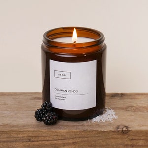 Pu&ograve; includere: Un barattolo di vetro marrone con una candela e un'etichetta bianca su cui &egrave; scritto "asha" e "OBI-WAN KENOBI". L'etichetta riporta anche "poured by hand" e "6oz soy candle". La candela &egrave; accesa e la fiamma &egrave; visibile.