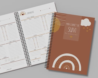 May include: A spiral-bound notebook with a terracotta cover featuring a white rainbow, a cloud, and the text Mon carnet de suivi. The open pages show daily tracking templates with sections for meals, naps, and notes, decorated with small rainbow icons.