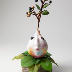 May include: A whimsical sculpture featuring a face-shaped form with a berry branch sprouting from the top. The face is painted with pastel colors and has black eyes and a small mouth. The sculpture rests on a wooden base with green leaves.