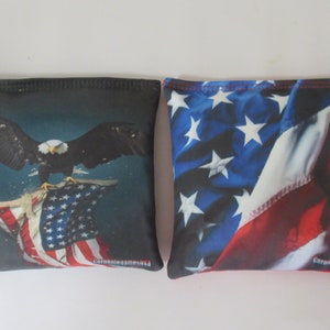 May include: Two bean bags with American flag designs. One bean bag features a bald eagle with outstretched wings landing on an American flag. The other bean bag features a close-up of an American flag. Both bean bags have the text "CornholeGameUSA" printed on the bottom.