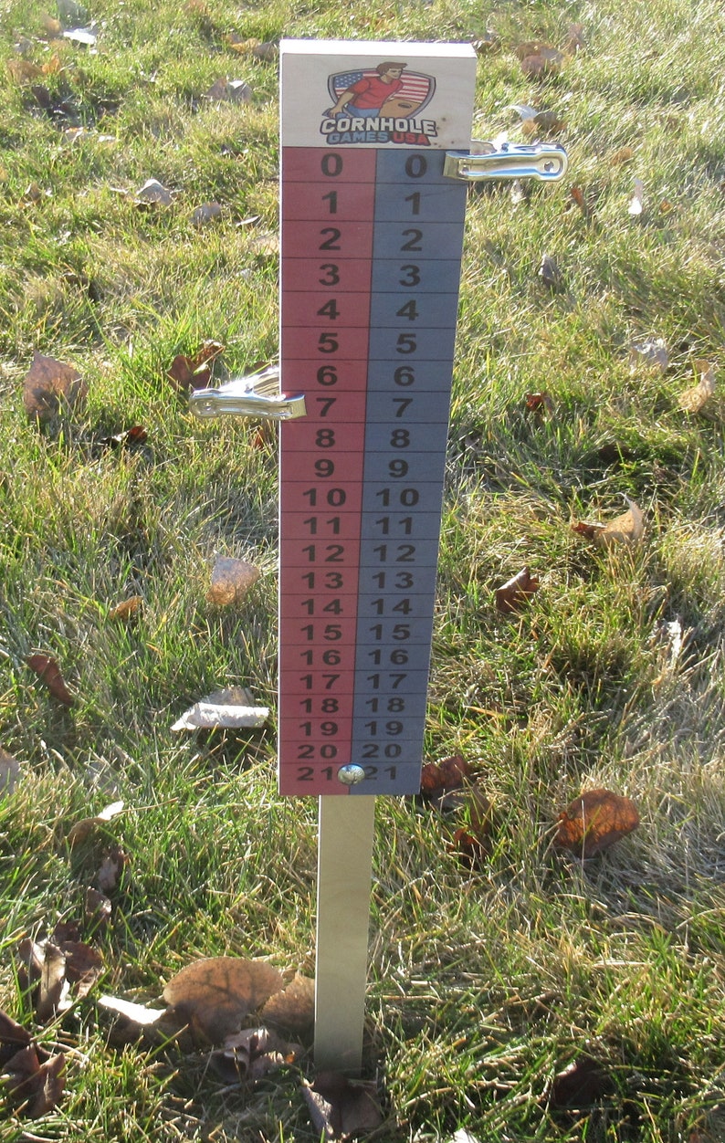 May include: A wooden cornhole scorekeeper with the text "CORNHOLE GAMES USA" and a graphic of a person throwing a bean bag. The scorekeeper has red and gray columns with numbers from 0 to 21. Metal clips are attached to the sides.