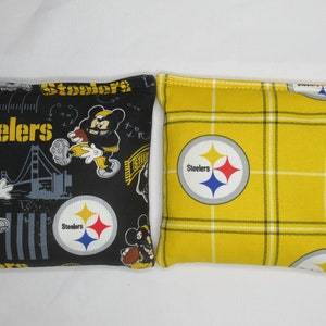 May include: Two black and yellow bean bags with the Pittsburgh Steelers logo. One bean bag has a black background with a Mickey Mouse design and the other has a yellow and black plaid pattern.