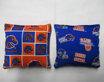 May include: Two blue and orange bean bags featuring the Boise State Broncos logo. One bean bag has a pattern of the Bronco horse logo and the other has a pattern of the Bronco horse logo, a football helmet, and a basketball.