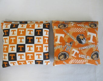 May include: Two orange and black bean bags with the University of Tennessee Volunteers logo. One bag has a checkered pattern with the letter "T" repeated, and the other bag has a houndstooth pattern with the words "Volunteers", "Tennessee", and "Vols" repeated.
