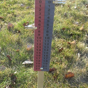 May include: A cornhole scorekeeper with a red and blue numbered display, ranging from 0 to 21. The top features the "Cornhole Games USA" logo. The scorekeeper is set in a grassy area.