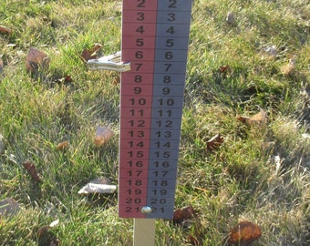 May include: A cornhole scorekeeper with a red and blue numbered display, ranging from 0 to 21. The top features the "Cornhole Games USA" logo. The scorekeeper is set in a grassy area.