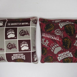 May include: Two bean bags with a maroon and gray pattern featuring the University of Montana Grizzlies logo, bear paw prints, and the words "Montana Grizzlies" and "The University of Montana".