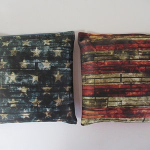 May include: Two fabric bean bags with an American flag design. The bean bags are made of a dark blue fabric with a distressed wood grain pattern. The stars are white and the stripes are red, white, and blue.