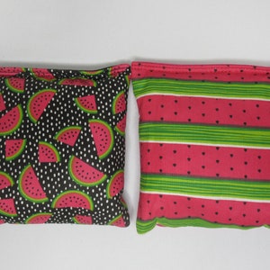 May include: Two fabric bean bags with watermelon patterns. One bean bag has a black background with white polka dots and red watermelon slices. The other bean bag has a red background with green stripes and small red watermelon seeds.