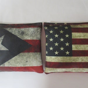 May include: Two bean bags with distressed flag designs. The left bean bag features the Puerto Rican flag with red, white, and blue stripes and a white star on a blue triangle. The right bean bag features the American flag with red and white stripes and a field of blue with white stars.