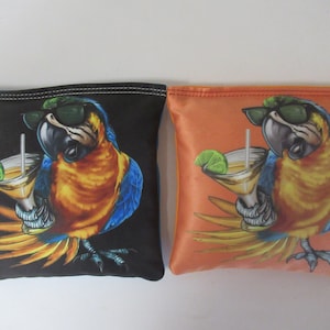 May include: Two square bean bags, one black and one orange, each featuring a cartoon parrot wearing sunglasses and holding a cocktail. The parrot has blue, yellow, and orange feathers. The cocktail has a lime wedge.