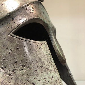 May include: A bronze Greek warrior helmet with a weathered patina. The helmet has a distinctive pointed crest and a face mask with a single eye opening.