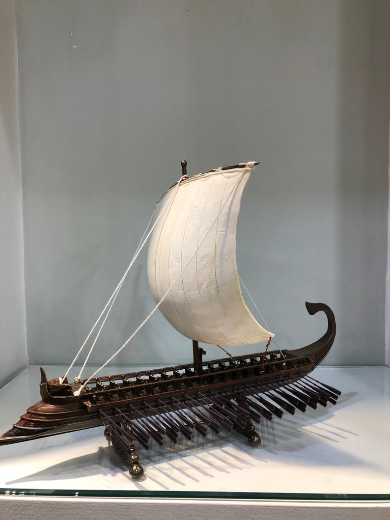 Puede incluir: Un modelo detallado de un antiguo barco de guerra trirreme griego con una vela blanca y un casco de madera marr&oacute;n. El barco tiene un dise&ntilde;o largo y estrecho con una proa puntiaguda y una popa curva. El modelo est&aacute; montado sobre una base de madera con una fila de remos a cada lado.